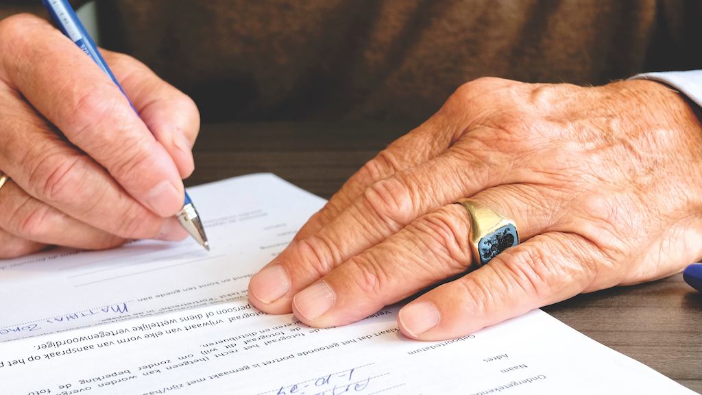  A close-up of a person's hands signing a contract. The pen is held in the right hand, while the left is holding the paper for stability. There is a ring on the left hand.