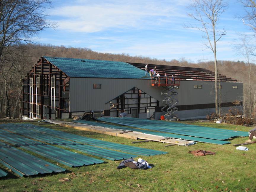 Metal building under construction. One wall is built, and half the roof has sheets. Other sheets are still on the ground.